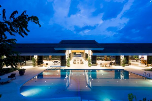 A luxurious modern building with a large illuminated swimming pool at night, surrounded by seating areas and lush greenery.