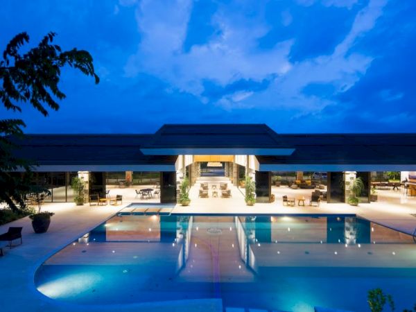A luxurious modern building with a large illuminated swimming pool at night, surrounded by seating areas and lush greenery.