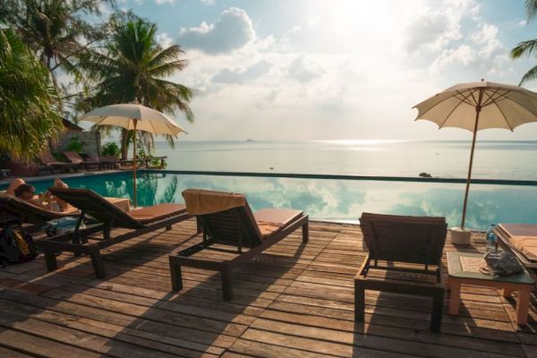 A serene poolside scene with lounge chairs, umbrellas, and palm trees overlooking a calm body of water under a bright sky.