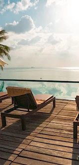 Deck chairs and umbrellas on a wooden deck by a pool with a view of the ocean, palm trees, and a bright sky.