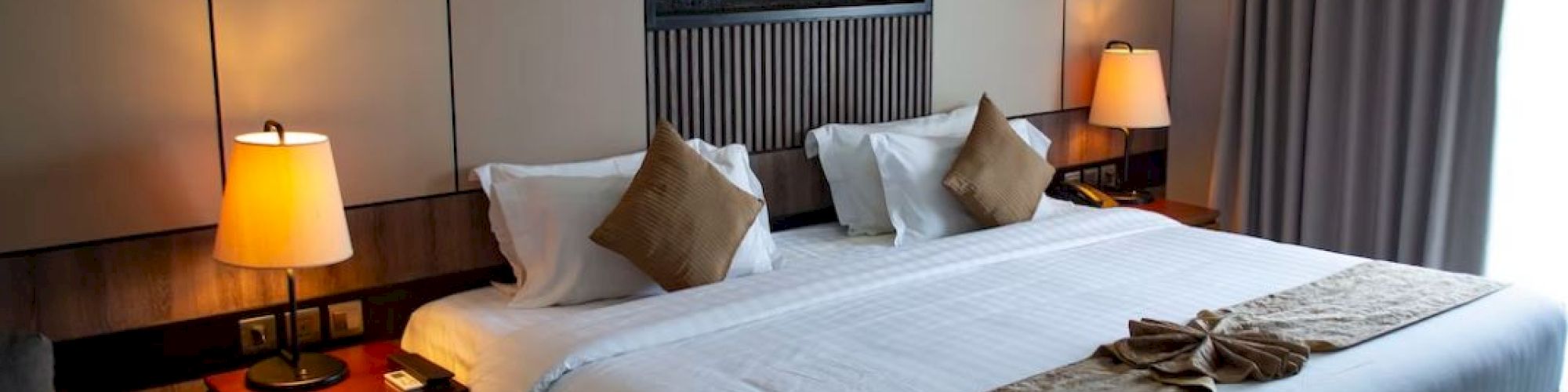 A hotel room with a large bed, brown pillows, two lamps, and a decorative piece above the headboard. Closed curtains are on the right.