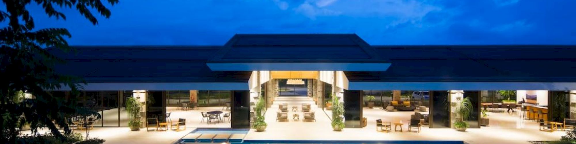 A modern building with open spaces overlooks a large, illuminated pool under a twilight sky, surrounded by seating and greenery.
