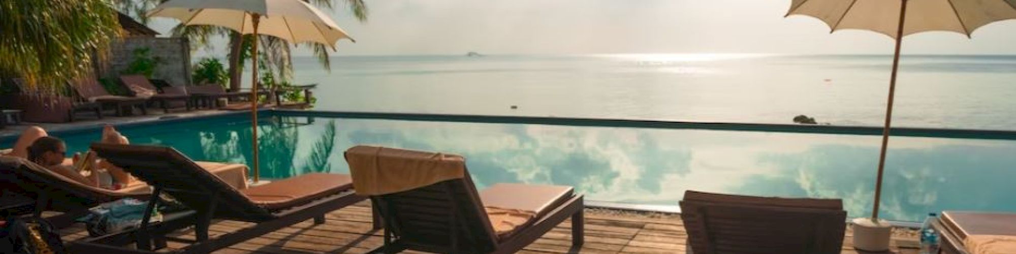 A serene swimming pool overlooking a tranquil ocean with lounge chairs and umbrellas, surrounded by palm trees under a partly cloudy sky.