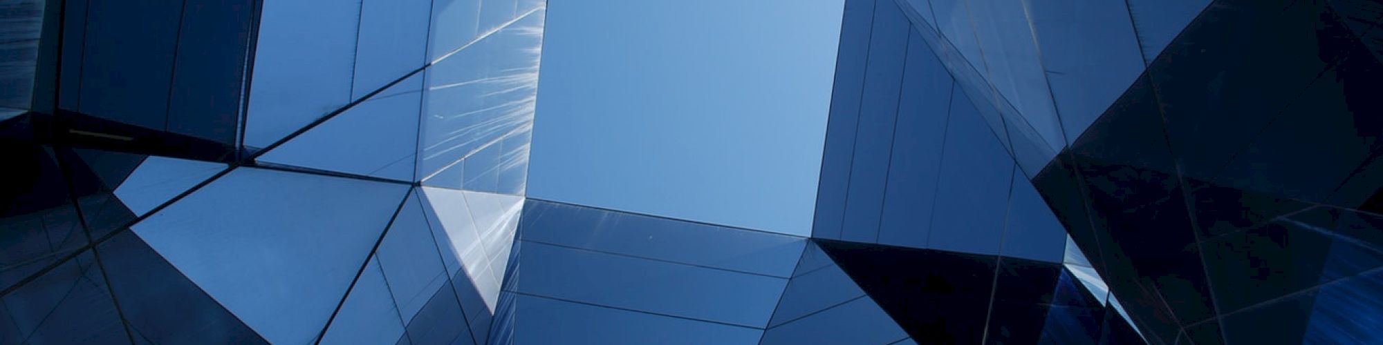 The image shows a geometric pattern of reflective building surfaces forming a skylight with a view of the blue sky.