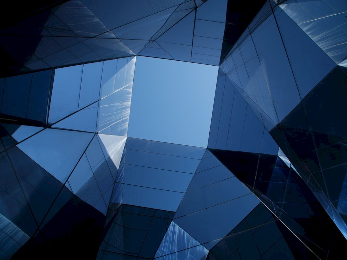 The image shows abstract, angular glass structures with reflections against the sky, creating a mirrored architectural effect.