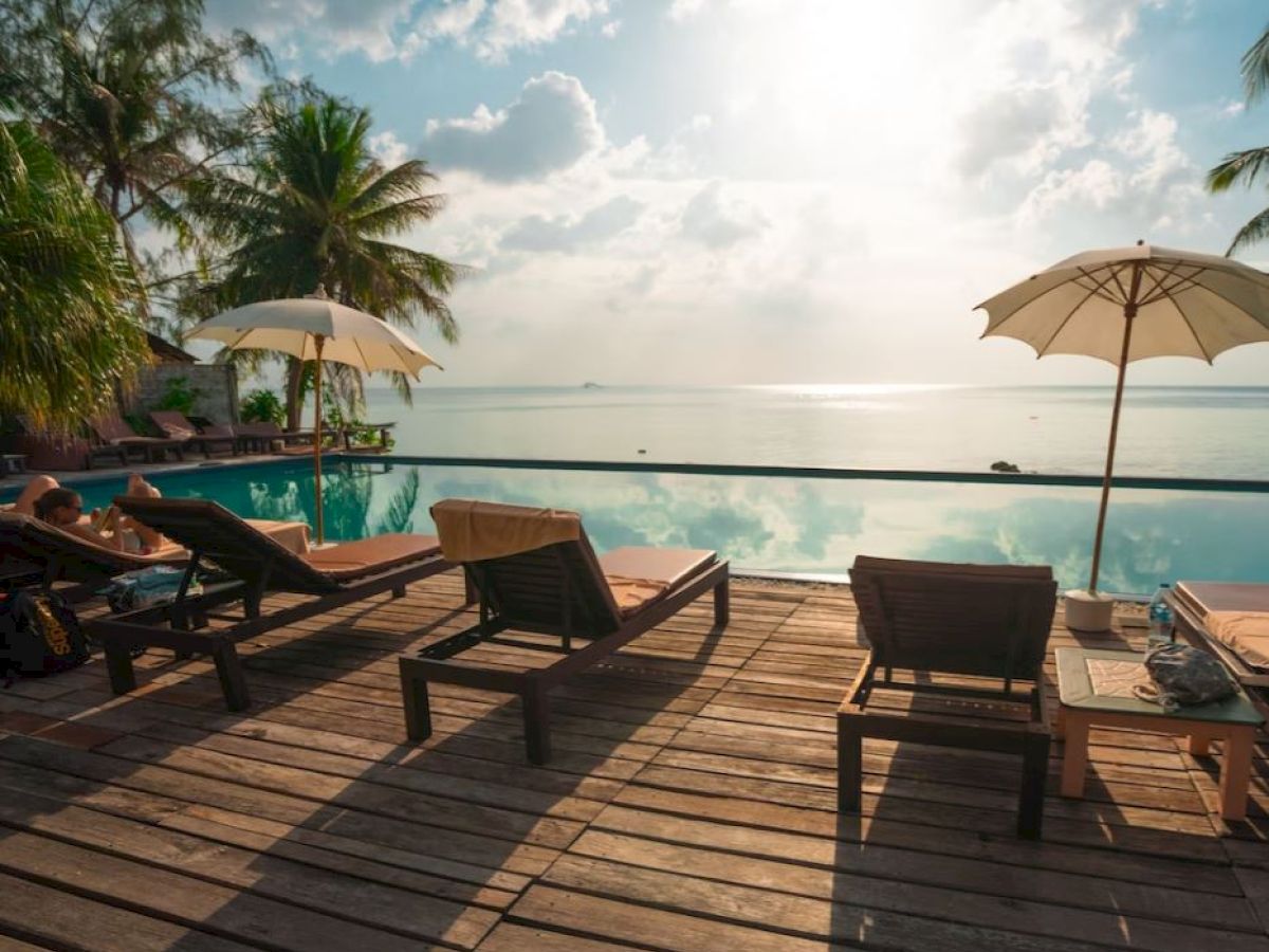 A tranquil poolside scene with lounge chairs, umbrellas, and palm trees overlooking a serene body of water under a sunny sky.