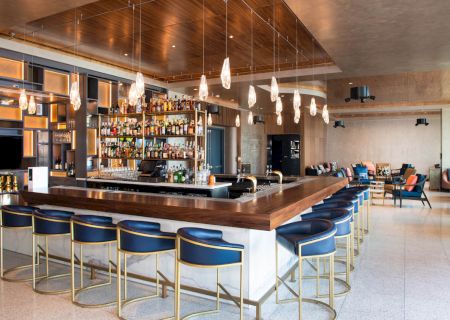 A modern bar with blue stools, a wooden counter, hanging lights, and shelves stocked with bottles, located in a spacious lounge area.