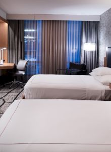 A modern hotel room with two beds, a TV, desk, and stylish decor. Warm lighting and a window with curtains add ambiance to the space.