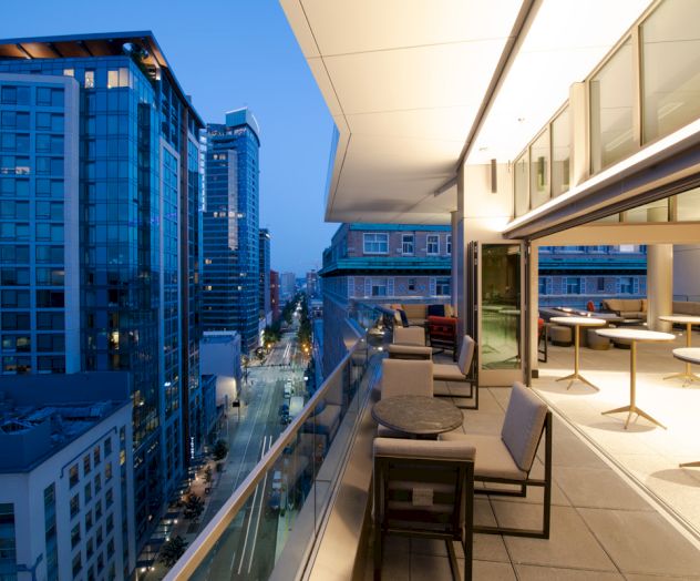 A modern balcony overlooking a cityscape at dusk, featuring outdoor seating and lighting, with tall buildings lining the street below.