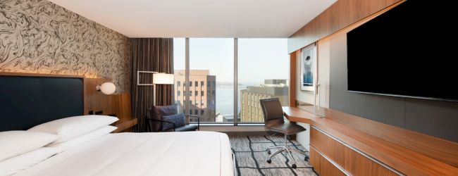 A modern hotel room features a large bed, a desk, a chair, and a flat-screen TV, with a window view of city buildings.