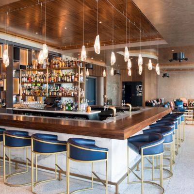 The image shows a modern bar with a wooden counter, shelves stocked with bottles, hanging lights, and blue chairs, in a spacious room.
