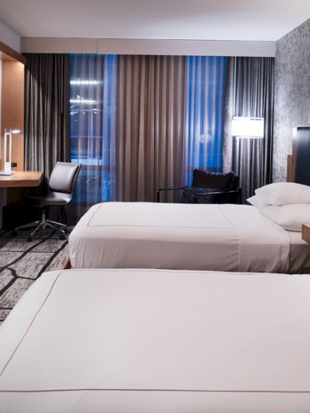 The image shows a modern hotel room with two beds, a TV, a desk, and stylish décor, illuminated by lamps and natural light.