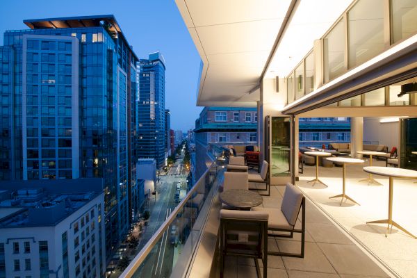 A modern rooftop terrace with tables and chairs overlooks a city street lined with tall buildings at dusk.