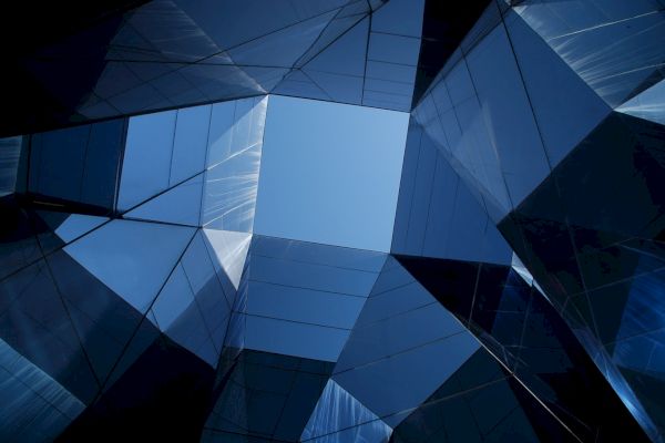The image shows modern, reflective buildings forming a geometric pattern, with a clear blue sky visible in the center.