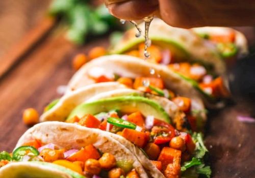 A hand squeezes lime over a row of tacos filled with chickpeas, diced vegetables, and avocado. Fresh lime slices are visible.