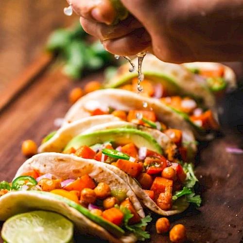 A hand squeezes lime over a row of tacos filled with chickpeas, diced vegetables, and avocado. Fresh lime slices are visible.
