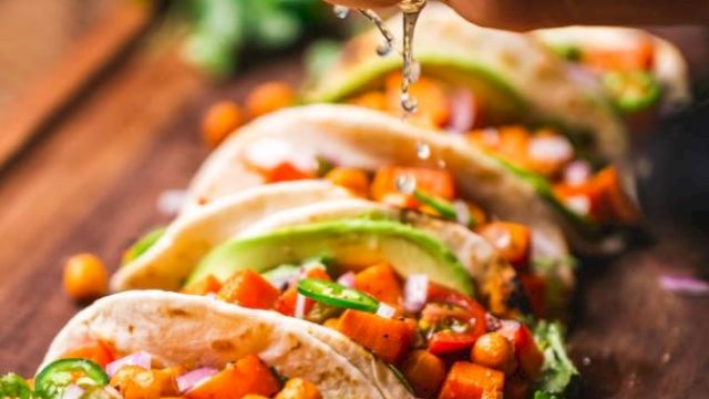 A hand squeezes lime over a row of tacos filled with chickpeas, diced vegetables, and avocado. Fresh lime slices are visible.