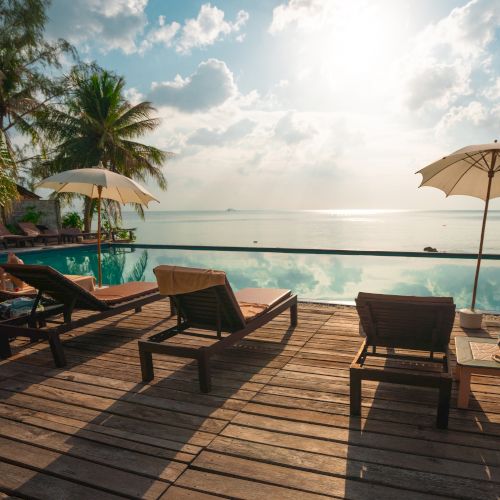 A serene beachfront view with lounge chairs, umbrellas, and trees by a calm body of water under a bright sky.