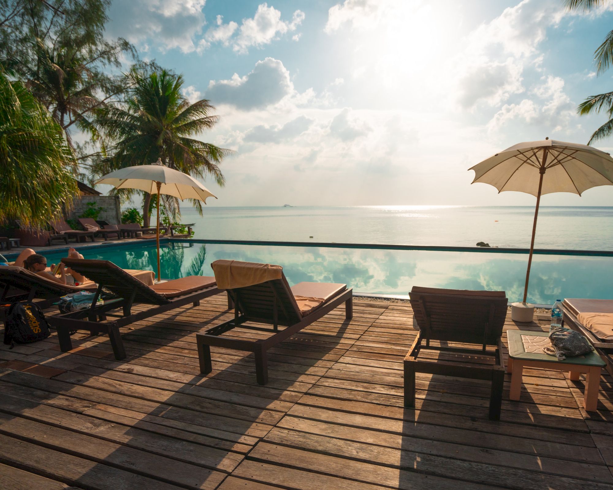 A tranquil scene with lounge chairs, umbrellas, a pool, and palm trees overlooking a serene ocean under a partly cloudy sky.