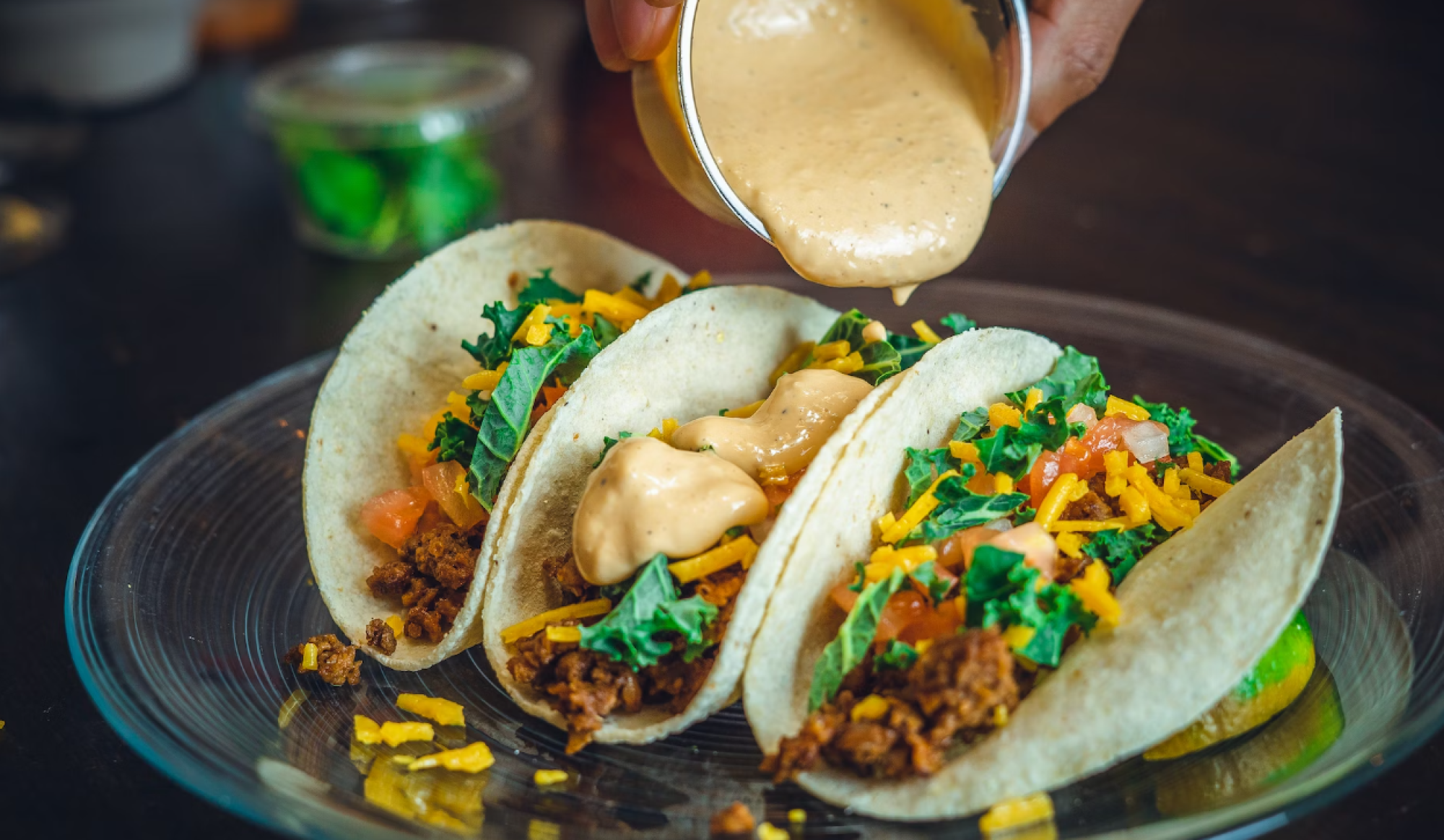 Three tacos filled with meat, lettuce, cheese, tomatoes being topped with a creamy sauce on a glass plate.