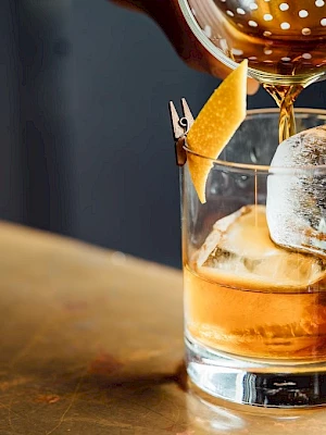 A cocktail being poured into a glass with ice and a twist of orange peel on the side.