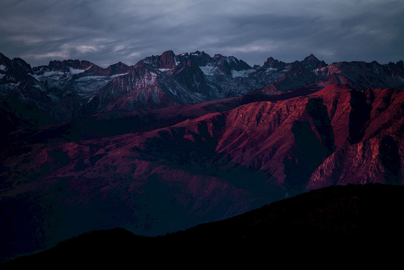 A dramatic mountain range at sunset, with rugged peaks and valleys bathed in a deep red and purple hue under a cloudy sky.