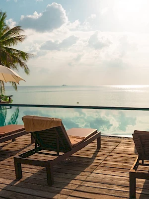 A serene poolside scene with lounge chairs, umbrellas, palm trees, and a calm ocean view under a partly cloudy sky completes the image.