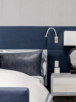 A modern bedroom with a navy and white theme, featuring a padded headboard, pillows, a bedside table with a lamp, and a wall-mounted light.
