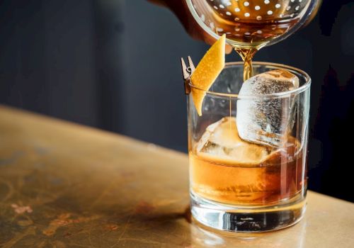 A cocktail being poured into a glass with ice and a citrus twist garnish on a bar surface.
