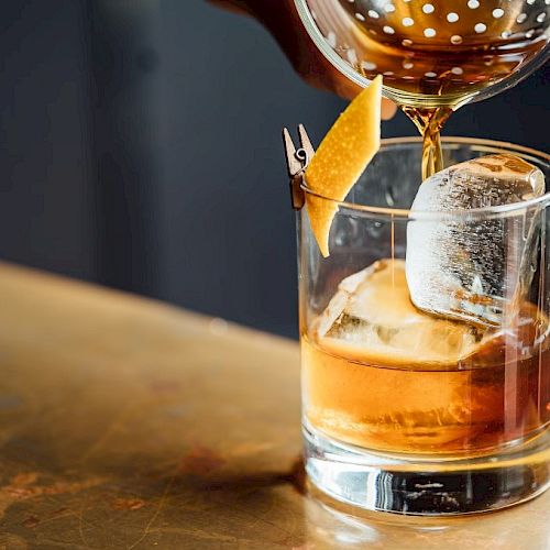 A cocktail being poured into a glass with ice and a citrus twist garnish on a bar surface.
