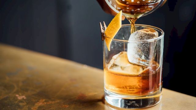 A cocktail being poured into a glass with ice and a citrus twist garnish on a bar surface.