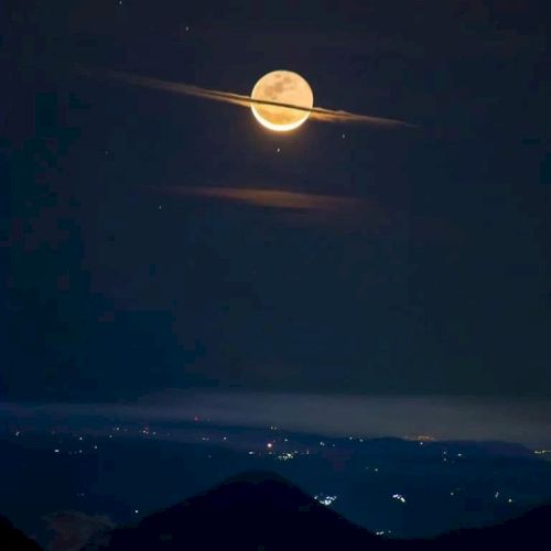 The image shows a full moon with clouds forming a ring around it, resembling Saturn, above a landscape with city lights scattered below.