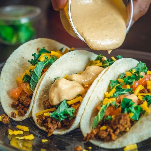 Three tacos filled with ground meat, cheese, kale, tomatoes, and sauce being poured. A small container of green items is in the background.