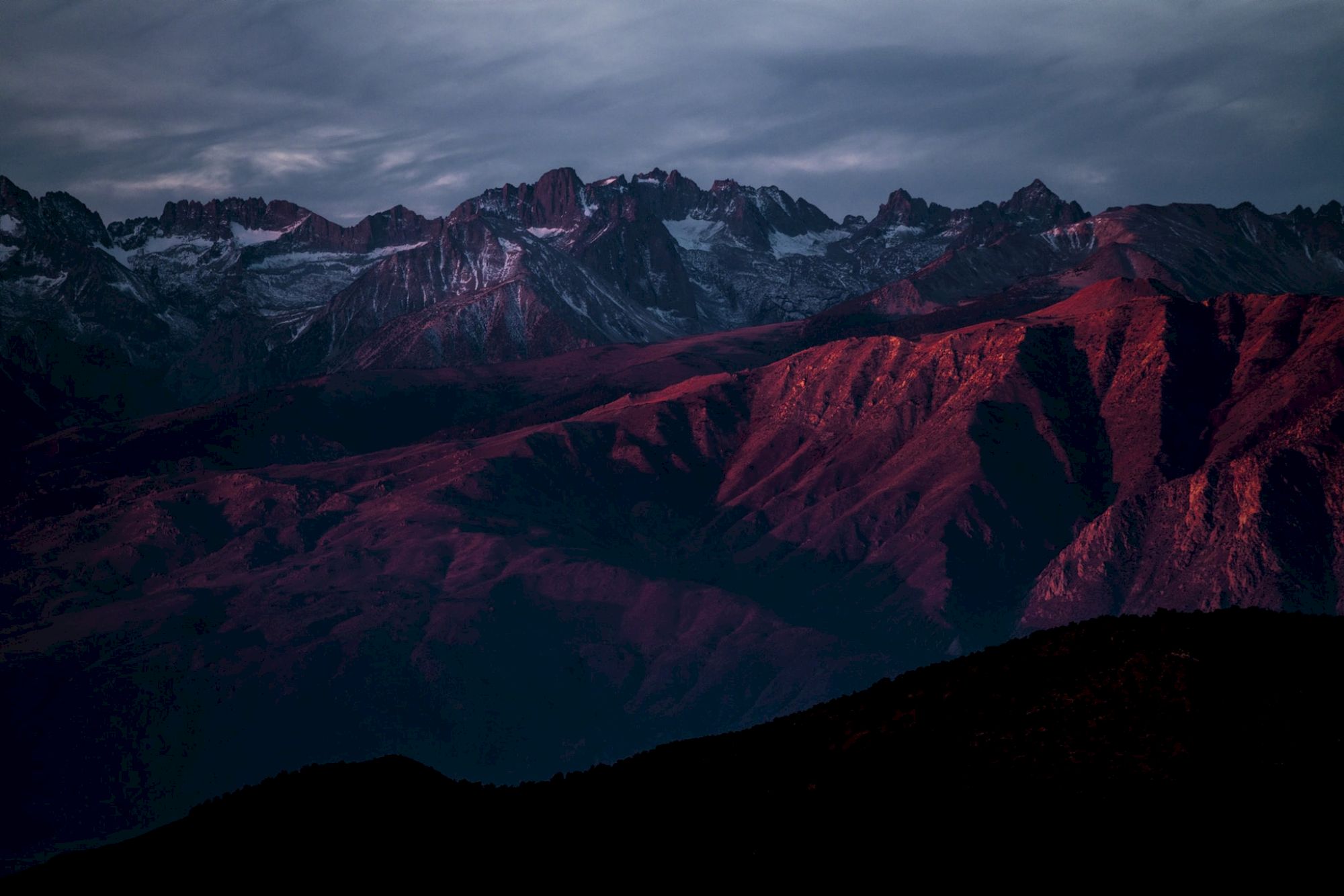 A mountain range with snow-capped peaks and red-hued lower slopes under a moody, cloudy sky at dusk or dawn.