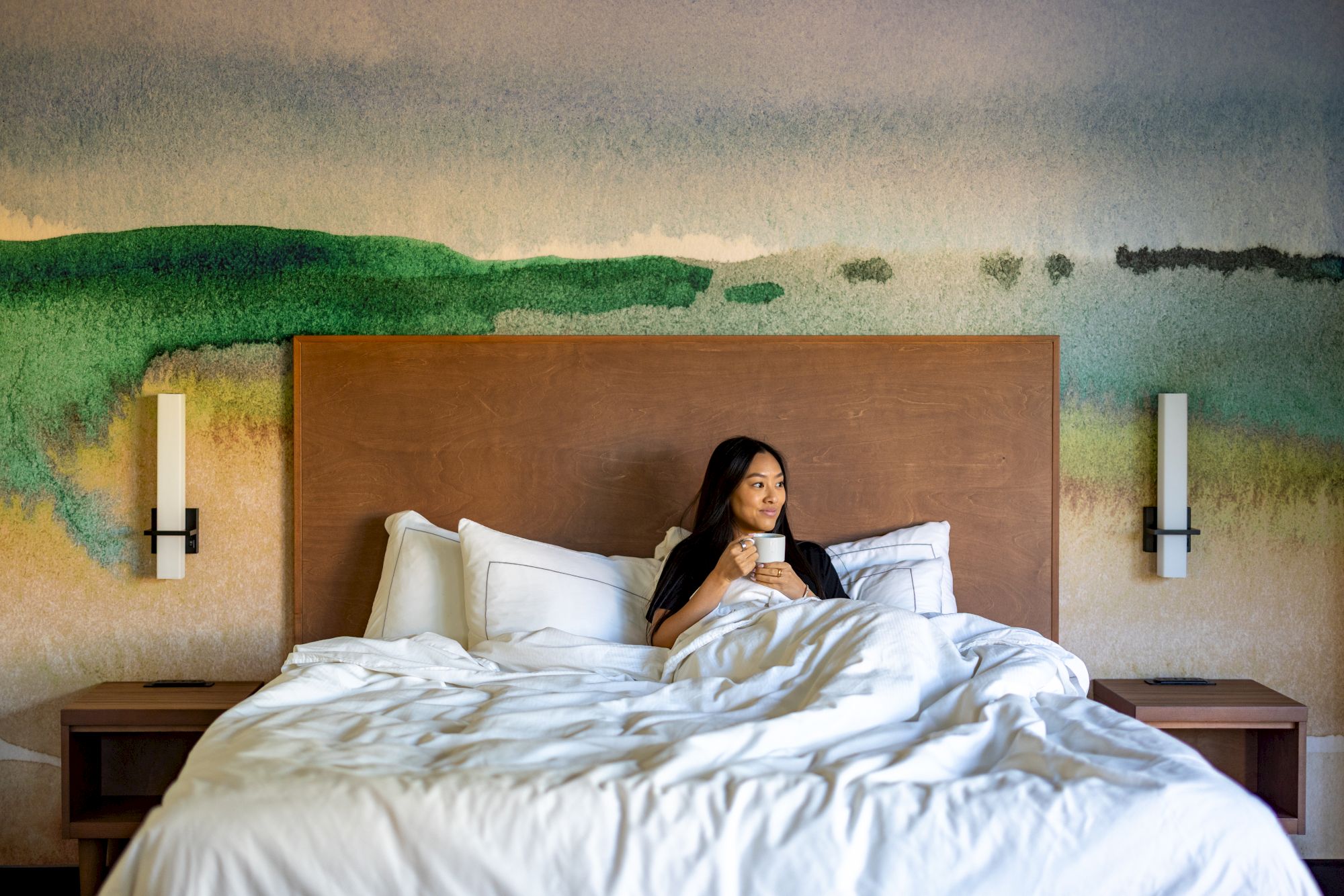 A person is lying in bed in a bright, neatly made hotel room with a mural wall behind, reading on a smartphone.