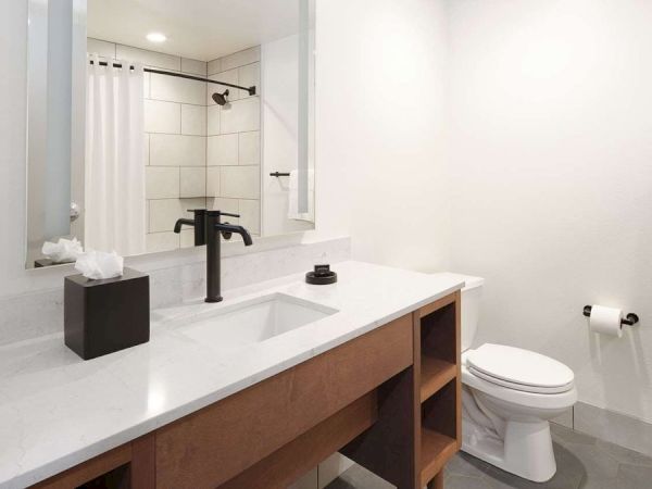 A modern bathroom with a white sink, dark faucet, toilet, shower, mirror, towels, and toiletries on a light-colored countertop.