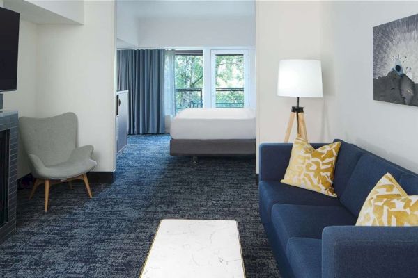 A cozy hotel suite features a fireplace, armchair, sofa with yellow pillows, and a bed by large windows with blue curtains.