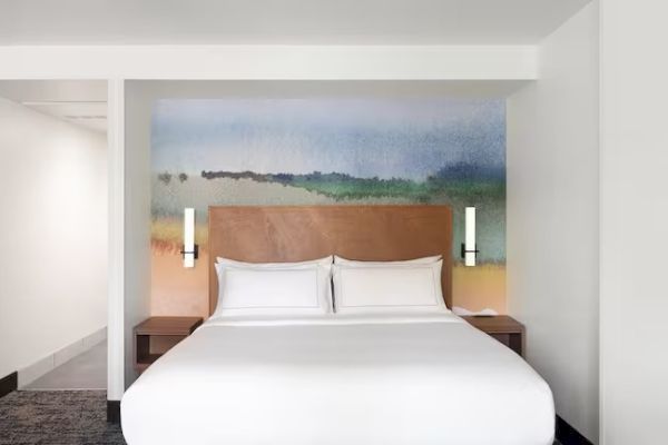 The image shows a modern hotel room with a bed, a colorful mural behind, and minimalistic decor on the walls.