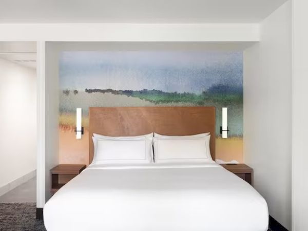 The image shows a modern hotel room with a bed, a colorful mural behind, and minimalistic decor on the walls.