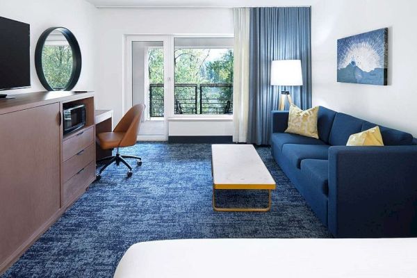 A modern hotel room features a bed, blue sofa, wooden desk with a chair, large TV, lamp, art on the wall, and a view outside the window.