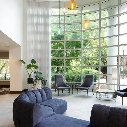 A modern living room with a large curved sofa, tall windows, and colorful hanging lights, overlooking a garden view through glass.