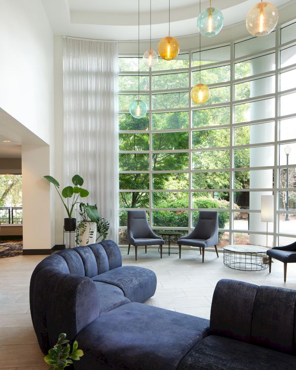 A modern living room with a large curved sofa, tall windows, and colorful hanging lights, overlooking a garden view through glass.