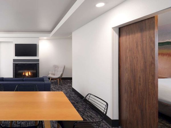 The image shows a modern living space with a fireplace, TV, sofa, dining table, and a glimpse of a bedroom through a sliding door.