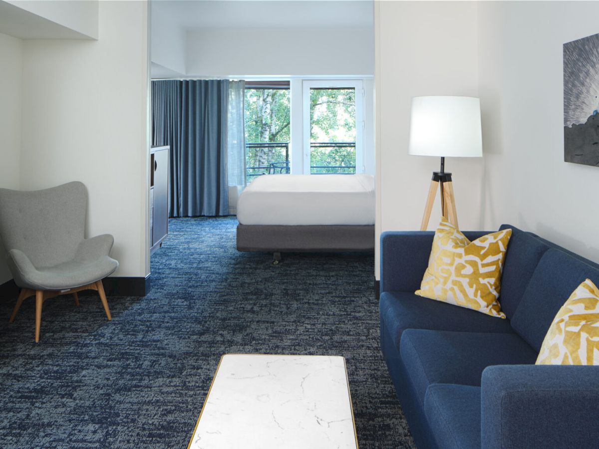 A modern hotel room featuring a blue sofa, fireplace, armchair, and a bed in the background.