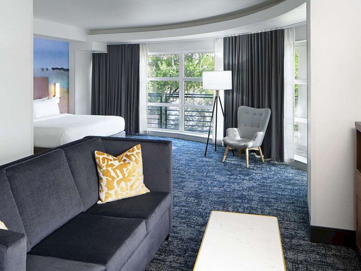 A hotel room with a bed, sofa, yellow pillows, a tall lamp, a grey armchair, and large windows with curtains.