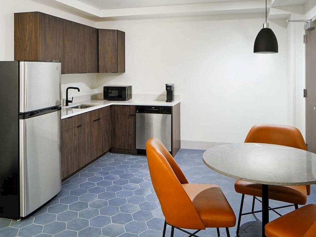 The image shows a modern kitchen with a fridge, oven, microwave, and coffee maker. There's a round table with orange chairs and hanging light.