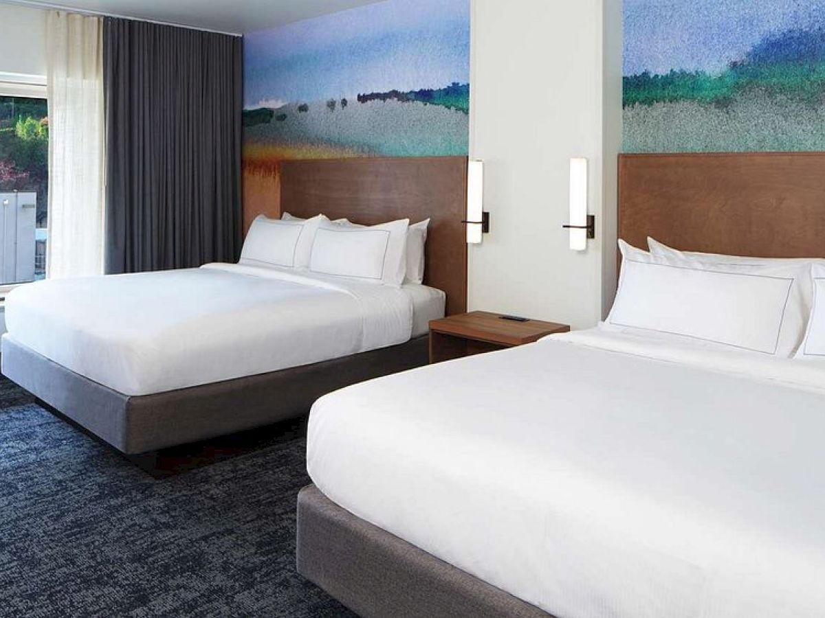 The image shows a hotel room with two large beds, modern decor, a colorful mural on the wall, and a window with a view outside.