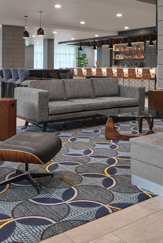 A modern lounge with a fireplace, gray sofas, patterned rug, and bar counter in the background, creating a cozy, stylish atmosphere.