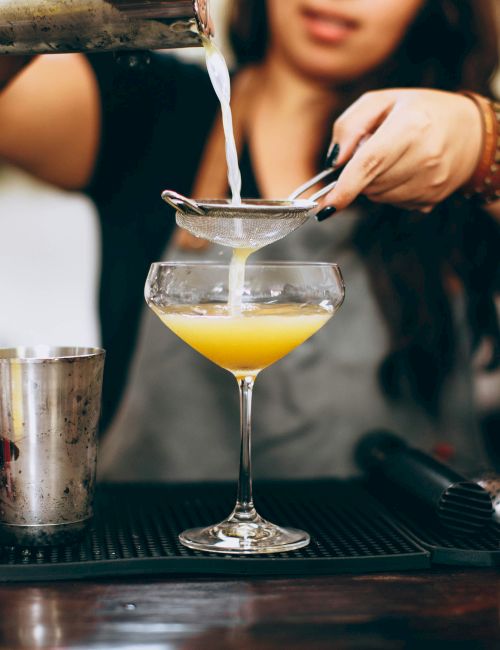 A person is straining a cocktail into a glass at a bar, surrounded by bottles and greenery.