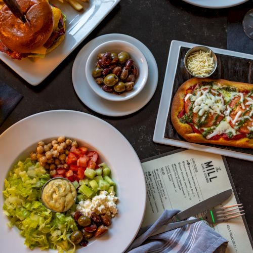 A colorful meal spread: a big salad bowl with greens, tomatoes, chickpeas, and toppings; a hot dog with toppings; a small bowl of olives; a plate with sliders and a beer; and a slice of bread with melted cheese, all on a dark table.
